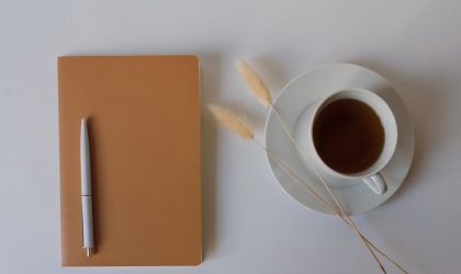 Reflexionsbuch Eine Tasse Kaffe und ein Notizbuch zur Reflexion liegen auf einem Tisch nebeneinander.