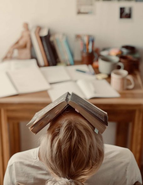 Chaos Eine Frau sitzt vor einem chaotischen Schreibtisch, hat ein offenes Buch über ihrem Gesicht liegen und wirkt überfordert und verunsichert.